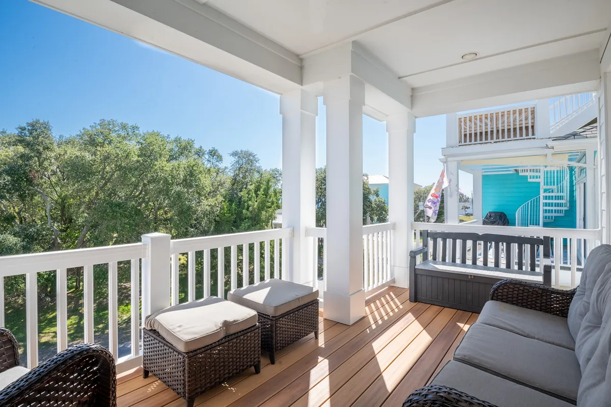 A serene wooden deck furnished with inviting wicker furniture, surrounded by lush green landscape and set beneath a bright blue sky. The setting is perfect for relaxation, socializing, or taking in the natural surroundings, with ample space for family and friends to gather outdoors.