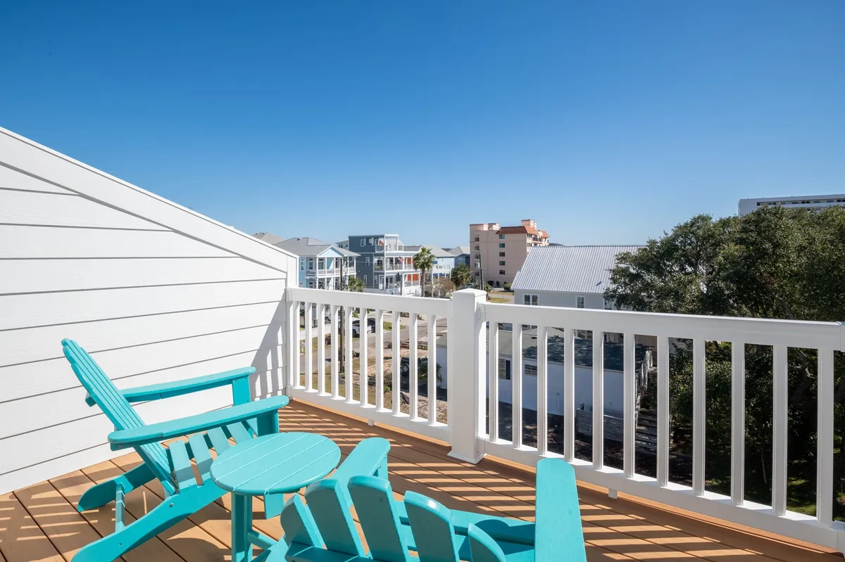 This exterior image highlights a balcony with turquoise chairs and a small round table, set against a backdrop of surrounding houses. The setup is tranquil, ideal for sipping coffee, reading, or enjoying the view, suggesting both sunrise and evening relaxation.