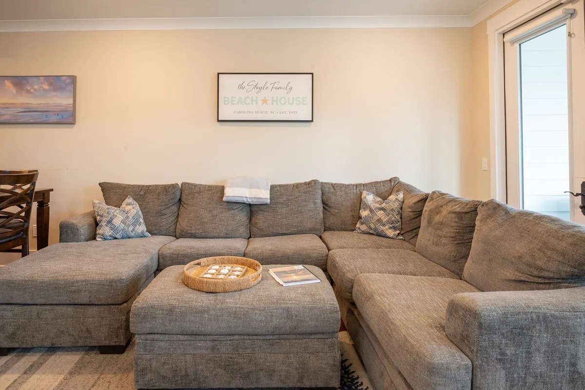 A spacious living area featuring a large sectional sofa, decorative pillows, a coffee table with coastal decor, a 'beach house' sign, and a doorway leading to the outside. The space feels inviting and homey, perfect for both people and pets to relax together after island adventures.