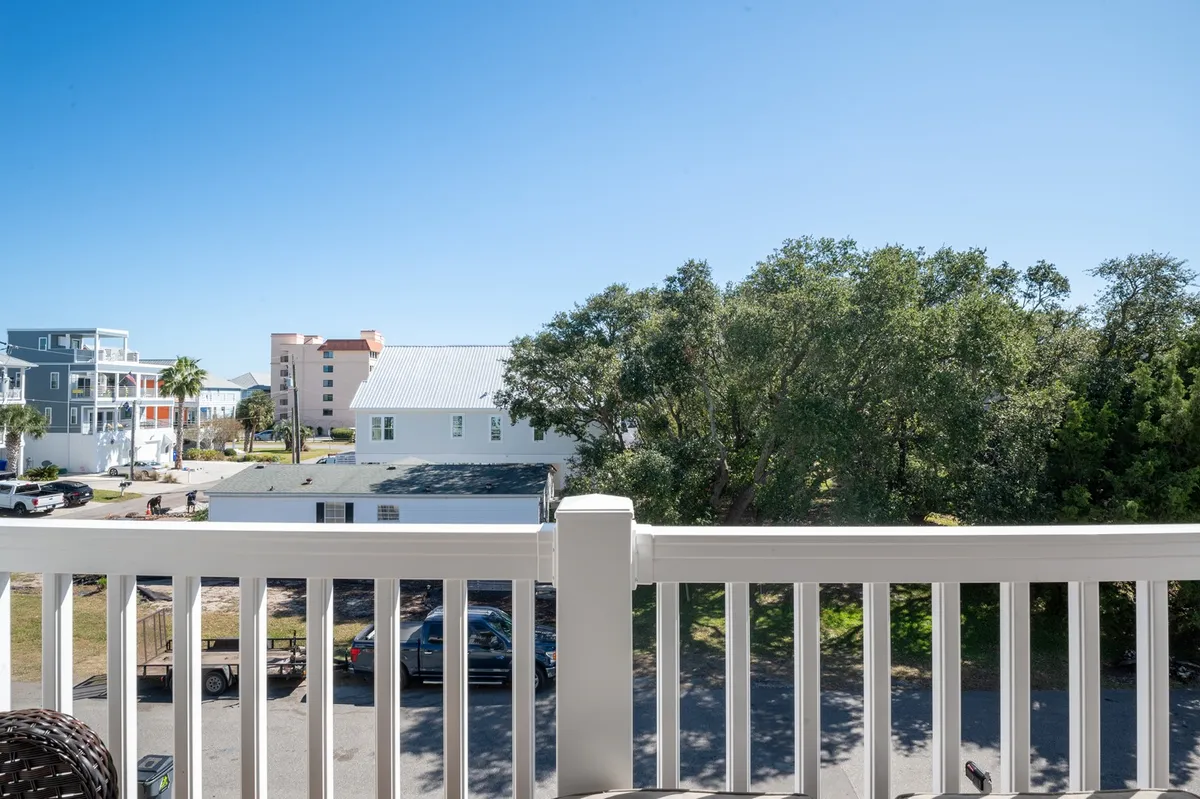 A peaceful balcony scene with a railing overlooking lush green trees and neighboring buildings. The light suggests either a tranquil morning or a golden sunset, inviting relaxation and outdoor enjoyment. The balcony serves as a transitional space between the home and the natural surroundings of the island.